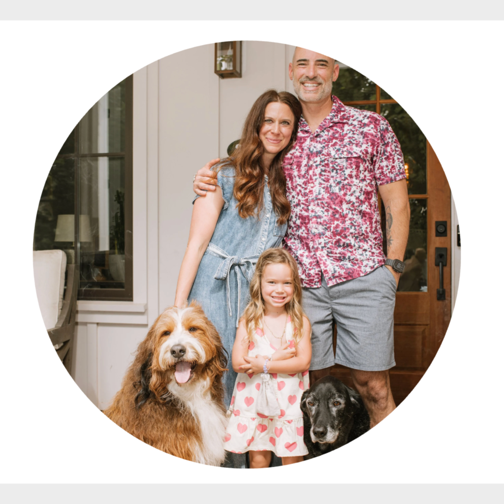 Family portrait of parents, young daughter, and two dogs standing together on a front porch outside their